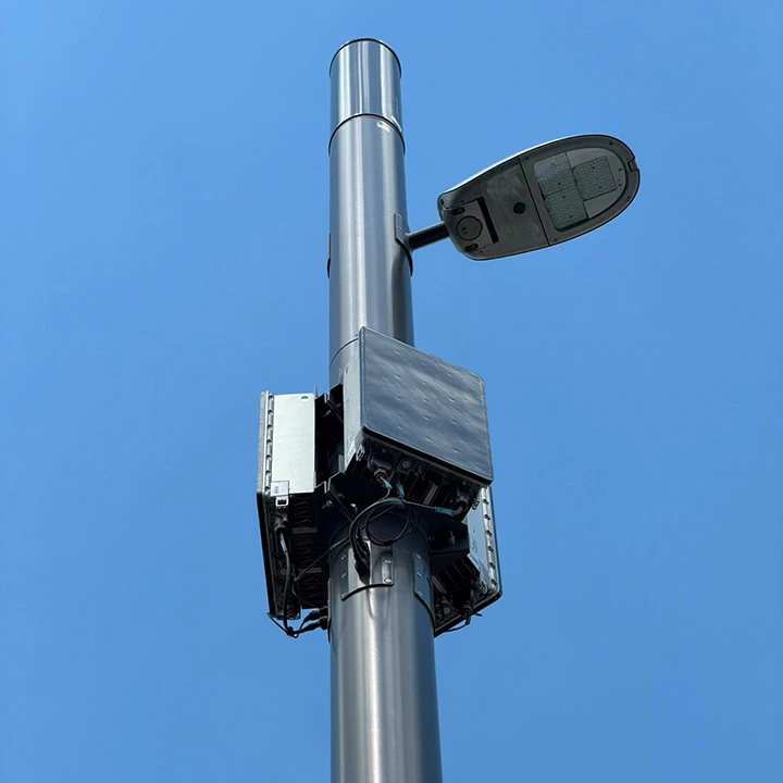 Nahaufnahme einer modernen Straßenlaterne, an deren Mast mehrere 5G-Antennen- und Technikmodule installiert sind. Die Aufnahme aus der Untersicht zeigt die Infrastruktur vor einem wolkenlosen blauen Himmel.