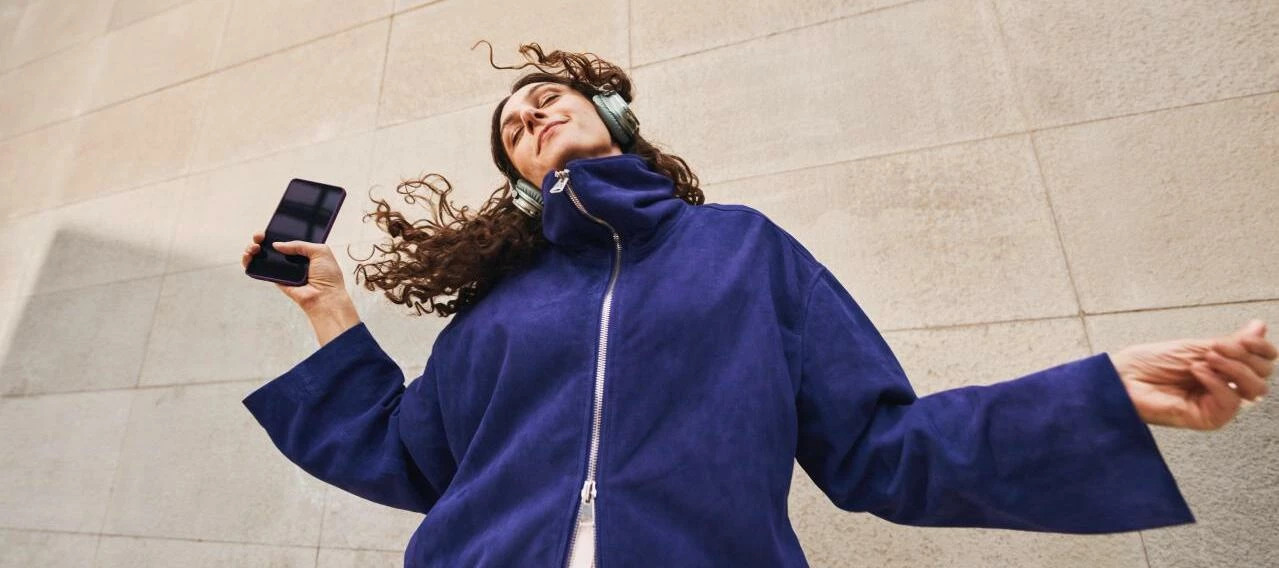 Eine Person mit großen Kopfhörern hört Musik, tanzt leicht und hält ein Smartphone in der Hand.
