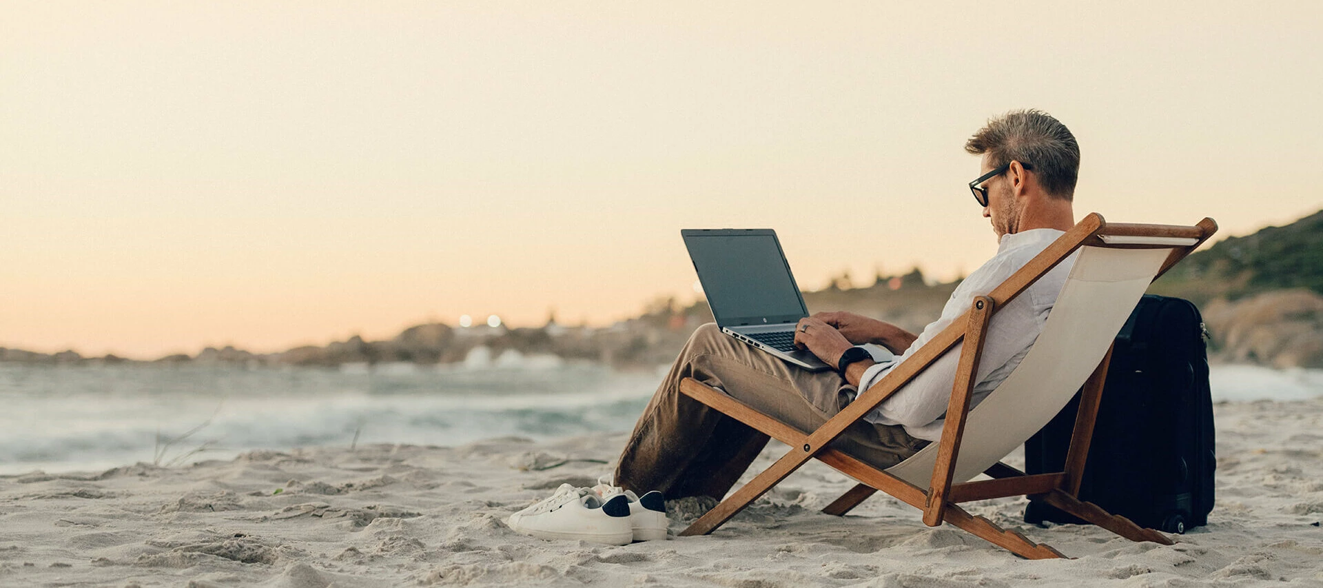En man på stranden i solstol, med dator och eftermiddagssol