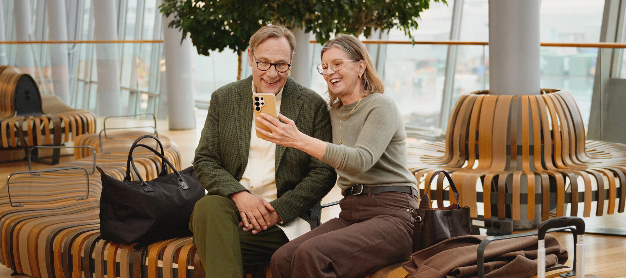 Två personer sitter på en bänk i en inomhusmiljö och tittar tillsammans på en mobiltelefon, med väskor placerade bredvid.