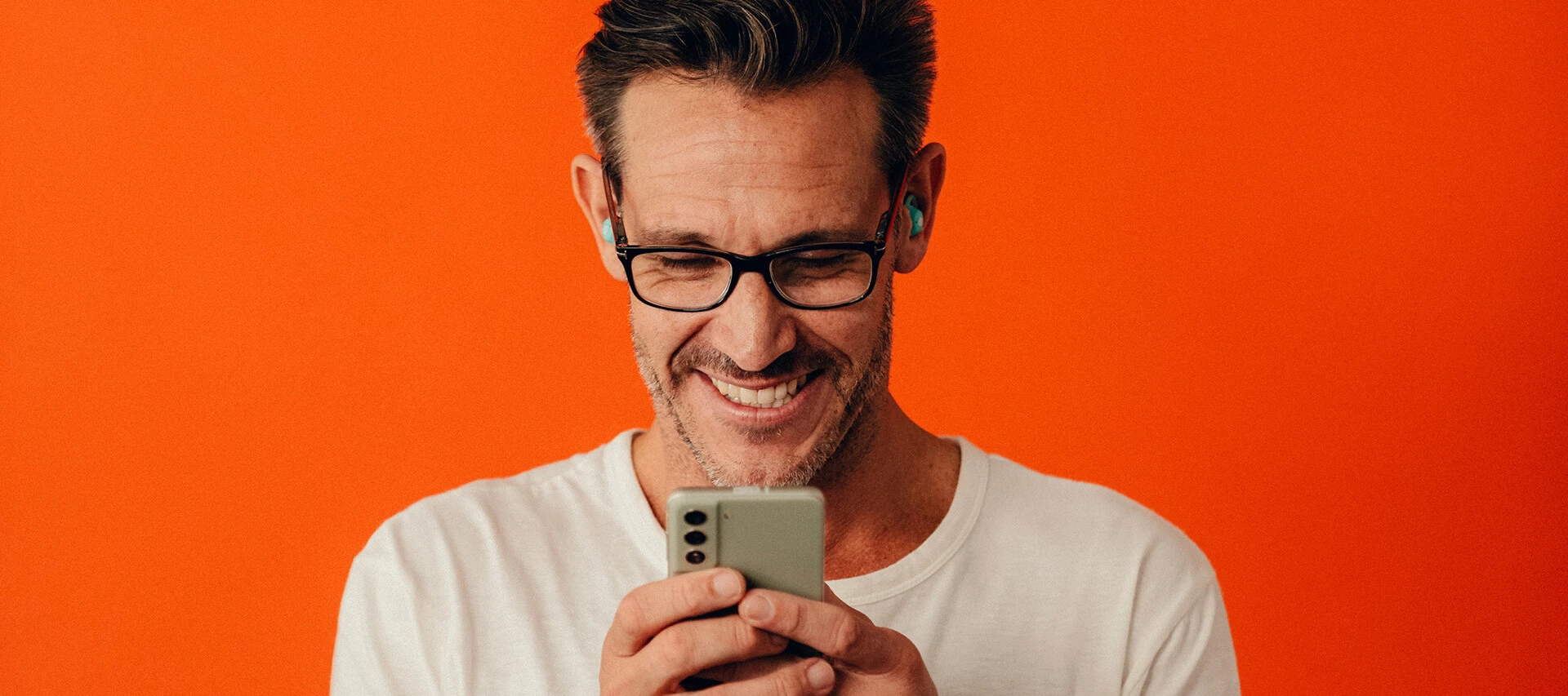 En leende man med glasögon tittar ner på sin mobiltelefon, med trådlösa hörlurar i öronen, mot en orange bakgrund.