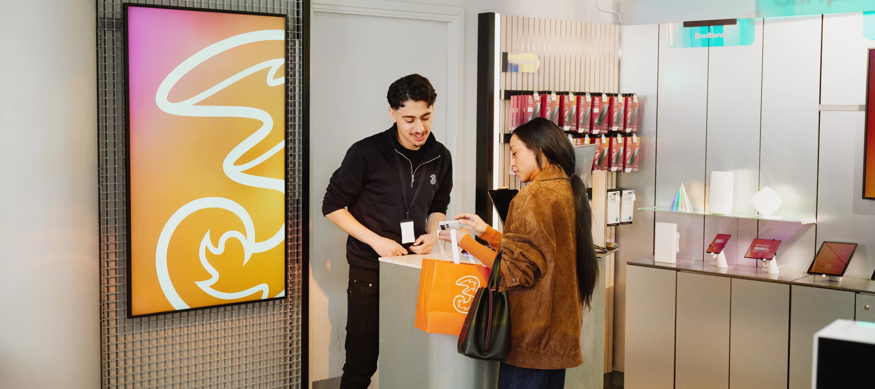 Två personer står vid en disk i en butik där Tre‑logotypen syns, och den ena räcker över en påse till den andra.