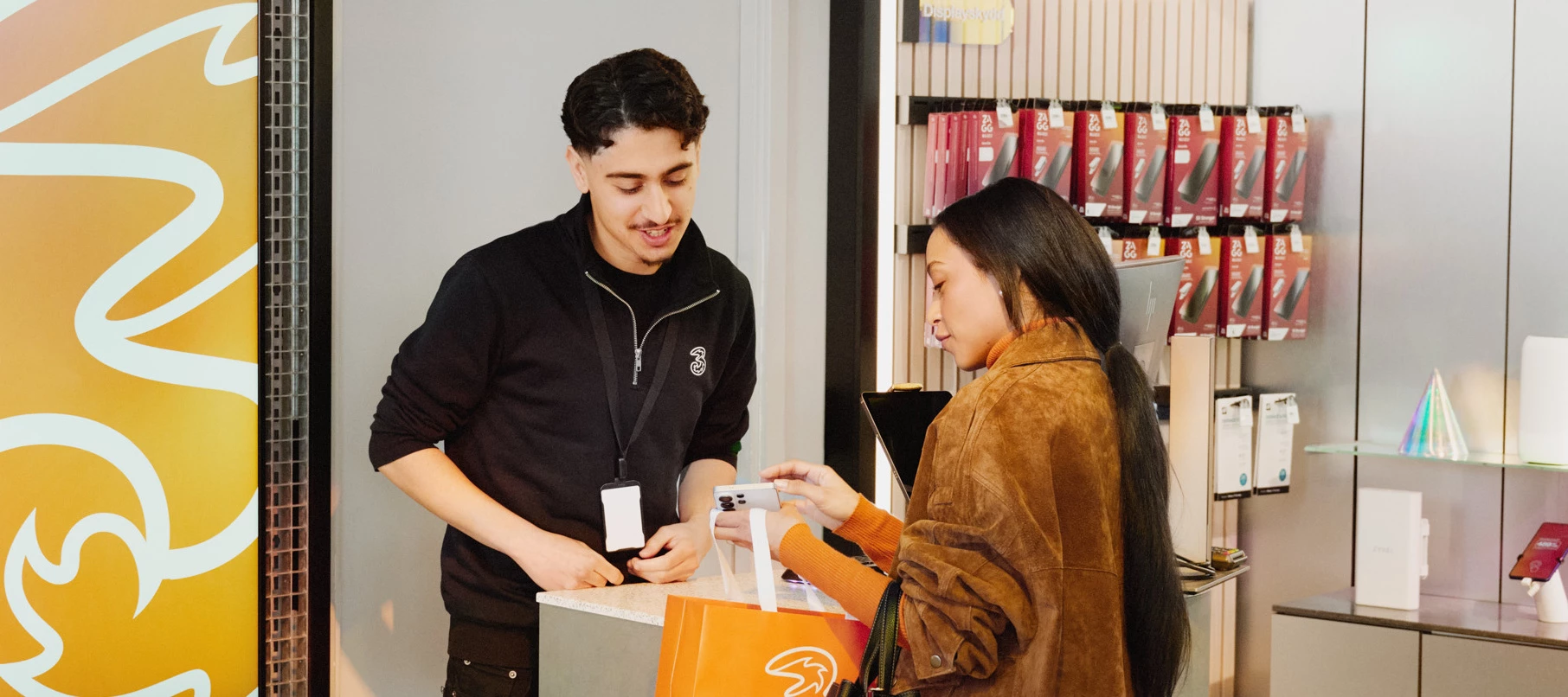 Två personer står vid en disk i en Tre‑butik, där en person räcker över en mobiltelefon.