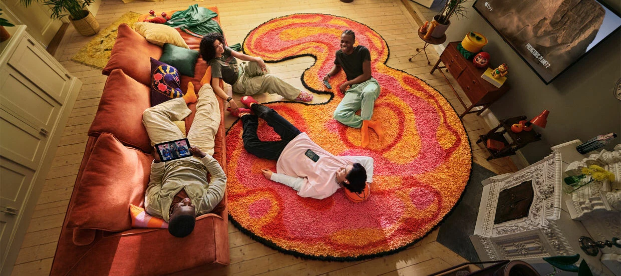 Fyra personer sitter och ligger avslappnat i ett vardagsrum på en stor färgglad matta formad som siffran 3.