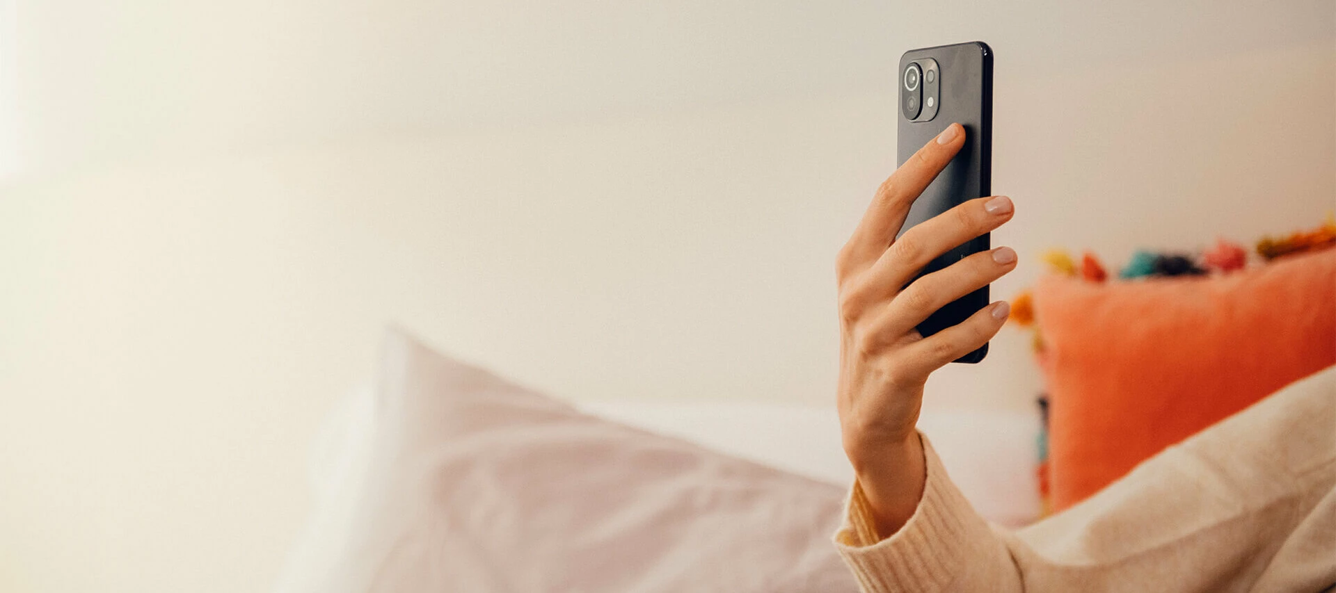 En person ligger på en säng och håller en smartphone i ena handen, med färgglada kuddar i bakgrunden.