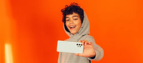 Ett barn i grå hoodie tar en selfie med en smartphone mot en orange bakgrund.